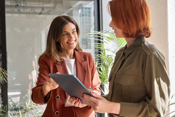 Zwei Frauen stehen in einem modernen Bürogebäude und unterhalten sich. Beide sind formell gekleidet und wirken konzentriert. Im Hintergrund sind große Fenster zu sehen, die viel natürliches Licht in den Raum lassen, sowie Büromöbel und Pflanzen, die eine professionelle Atmosphäre schaffen.