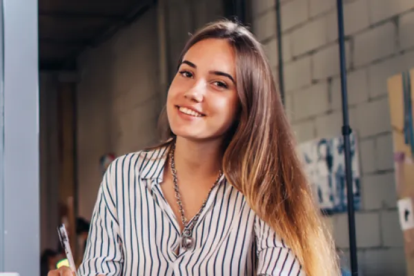 Eine junge Frau sitzt an einem Schreibtisch mit einem Notizbuch und lächelt freundlich in die Kamera. Sie trägt ein gestreiftes Hemd, und im Hintergrund ist eine kreative Büroumgebung zu sehen. Der Schreibtisch ist mit Laptop, Notizen und einer Kaffeetasse bedeckt, während sie einen Stift in der Hand hält, bereit zu schreiben.