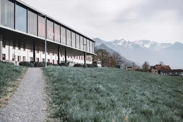 Campus Liechtenstein mit Glasfassade, Wiesenweg und Panoramablick auf die umliegenden Berge und das Dorf Vaduz.