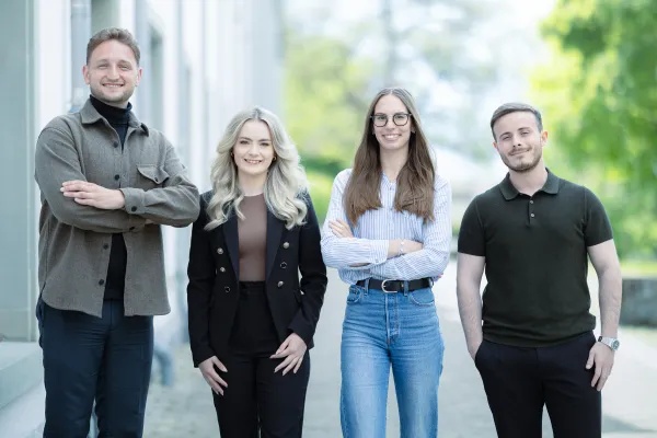 Vier Studierende im Masterstudiengang Information Systems der Universität Liechtenstein stehen nebeneinander auf einem Gehweg vor einem Universitätsgebäude und lächeln in die Kamera. Die Kleidung reicht von Business- bis Casual-Stil.