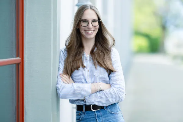 Eine weibliche Studierende im Masterstudiengang Information Systems trägt ein hellblau-weiß gestreiftes Hemd und Jeans, steht mit verschränkten Armen an einer Wand und lächelt offen.