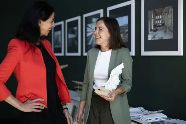 Zwei Frauen im Gespräch vor einer Bilderwand an der Universität Liechtenstein, eine hält ein Architekturmodell.