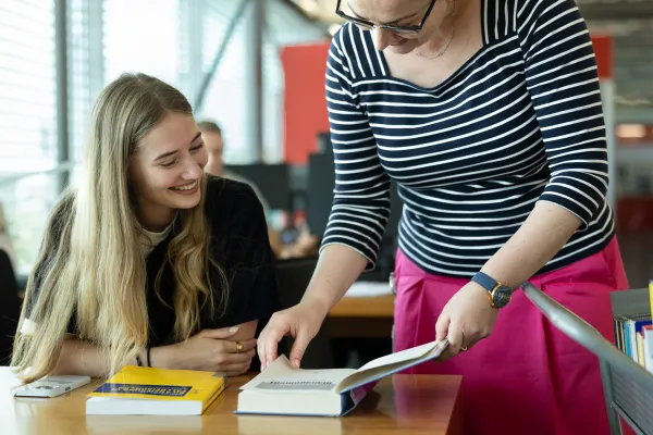Eine Bibliotheksmitarbeiterin zeigt einer lächelnden Studentin Inhalte aus einem geöffneten Buch.