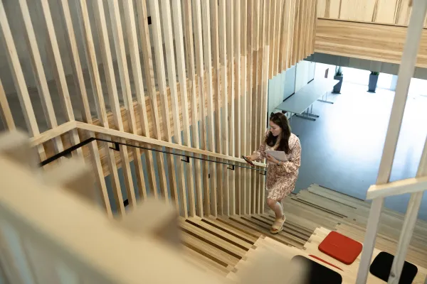 Studentin steigt eine moderne Holztreppe in der Universität Liechtenstein hinauf und schaut auf ihr Smartphone.