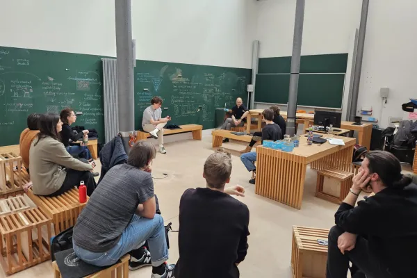 Eine Gruppe von Studierenden sitzt in einem modernen Seminarraum der Universität Liechtenstein, umgeben von Tafeln mit Notizen und Skizzen. Sie diskutieren angeregt, während ein Dozent vorne sitzt und zuhört.