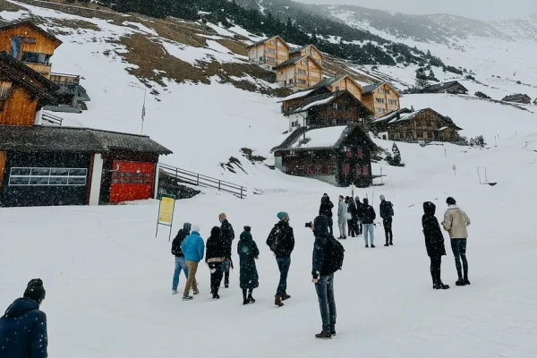 Eine Gruppe von Menschen steht in einer verschneiten Berglandschaft vor traditionellen Holzhäusern. Im Hintergrund sind schneebedeckte Hügel und Nadelbäume zu sehen. Die Szene vermittelt eine winterliche Atmosphäre und zeigt eine Verbindung von Architektur und Natur.