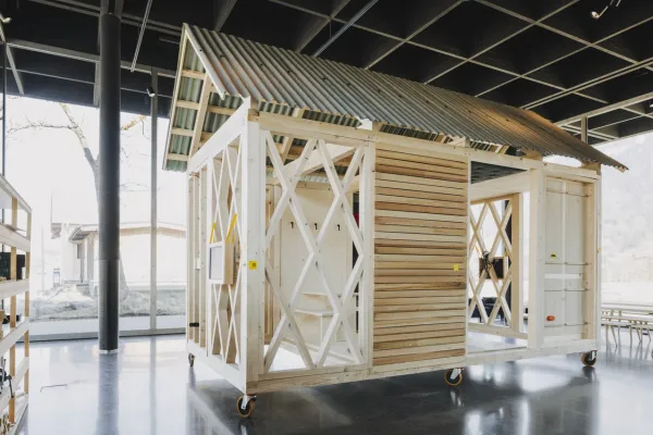 Ein einfaches, modulares Holzgebäude mit einem Wellblechdach steht auf Rollen in einem modernen, lichtdurchfluteten Raum. Das Design zeigt eine offene Struktur mit diagonalen Verstrebungen und Holzverkleidung, ideal für nachhaltiges Bauen und architektonische Bildung.