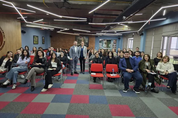 Seminarraum mit Studierenden und Dozenten, die an einem Cybercrime-Seminar teilnehmen. Die Teilnehmenden sitzen auf roten Stühlen in einem modernen Raum mit geometrischem Teppichmuster und futuristischer Beleuchtung.