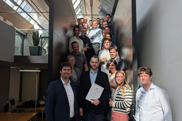 Eine Gruppe von Menschen steht auf einer Treppe in einem modernen Gebäude. Im Vordergrund hält eine Person ein Dokument in der Hand, umgeben von lächelnden Kollegen. Die Szene ist gut beleuchtet durch ein Oberlicht.