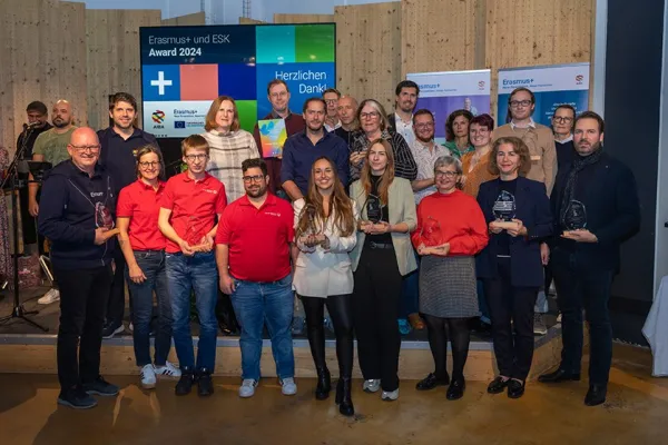 Eine Gruppe von Menschen steht auf einer Bühne und hält Trophäen. Im Hintergrund sind Banner mit Erasmus+ Logos und ein Bildschirm mit der Aufschrift "Erasmus+ und ESK Award 2024" zu sehen. Die Personen lächeln und posieren für ein Gruppenfoto.