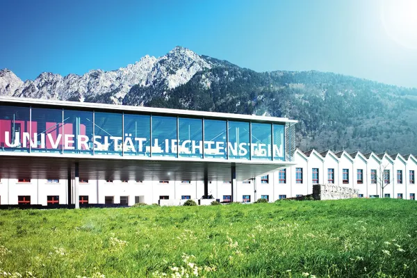 Außenansicht der Universität Liechtenstein mit modernem Glasgebäude vor malerischer Bergkulisse, umgeben von grüner Wiese.