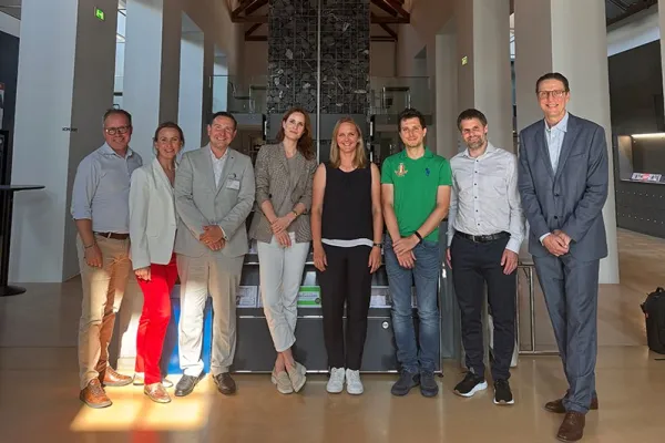 Eine Gruppe von neun Personen steht in einer modernen Halle mit Holzdach und einem Kronleuchter. Sie lächeln in die Kamera. Im Hintergrund sind hohe Fenster und eine Steinwand zu sehen.