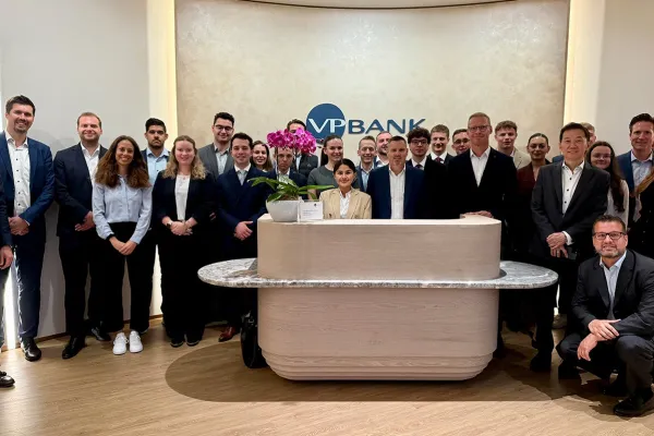 Gruppe von Studierenden und Fachleuten in formeller Kleidung steht in der Lobby der VP Bank. Im Hintergrund ist das Logo der Bank sichtbar. Die Szene vermittelt eine professionelle und lernorientierte Atmosphäre.