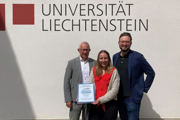 Drei Personen stehen vor einer Wand mit dem Logo der Universität Liechtenstein. Eine Person hält eine Auszeichnung in den Händen.