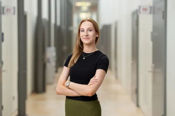 Eine junge Frau steht selbstbewusst mit verschränkten Armen in einem modernen Flur. Sie trägt ein schwarzes Oberteil und einen grünen Rock.