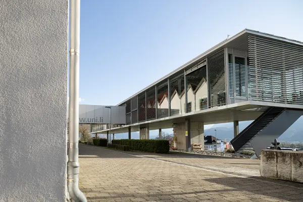 Modernes Gebäude der Universität Liechtenstein mit verglaster Fassade und Außentreppe, umgeben von einer gepflasterten Fläche und Hecken, vor einem klaren blauen Himmel.