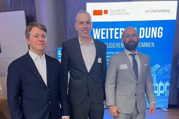 Drei Männer in Anzügen stehen vor einem Schild der Universität Liechtenstein. Im Hintergrund ist ein Banner mit der Aufschrift "Weiterbildung" zu sehen.