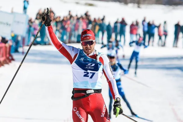 Langläufer überquert bei sonnigem Winterwetter als Sieger die Ziellinie eines Langlaufrennens, trägt einen rot-weißen Rennanzug mit Startnummer 27, hebt den Arm zum Jubel – im Hintergrund jubelnde Zuschauer und schneebedeckte Landschaft.