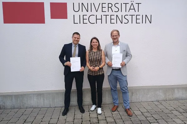 Drei Personen stehen vor einer Wand mit dem Logo der Universität Liechtenstein. Zwei von ihnen halten Zertifikate in den Händen.