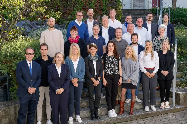 Eine Gruppe von Studierenden und Dozenten des LL.M.-Executive-Masterstudiengangs im Gesellschafts-, Stiftungs- und Trustrecht der Universität Liechtenstein posiert auf einer Treppe im Freien. Sie sind Teil des I&F Family Wealth Preservation Awards. Im Hintergrund sind Pflanzen und eine Gebäudefassade zu sehen.