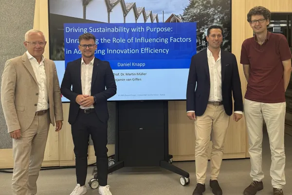 Vier Männer stehen vor einem Bildschirm, der eine Präsentation mit dem Titel „Driving Sustainability with Purpose“ zeigt. Sie tragen Business-Kleidung und lächeln in die Kamera. Der Hintergrund ist ein moderner Innenraum.