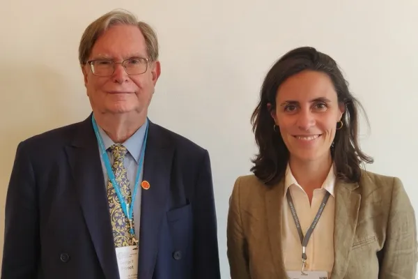 Zwei Personen stehen nebeneinander und lächeln in die Kamera. Beide tragen Namensschilder und sind formell gekleidet.