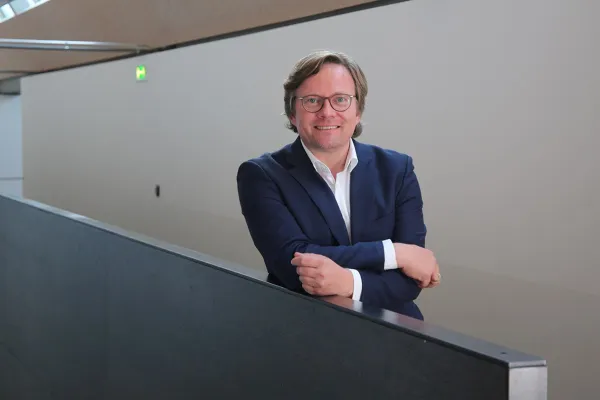 Ein Mann in einem blauen Anzug lehnt lächelnd an einem Geländer in einem modernen Gebäude.