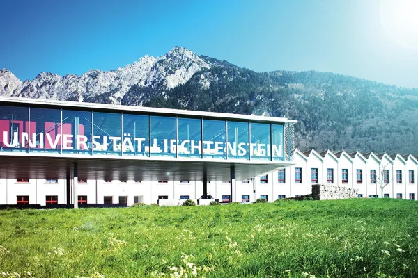 Außenansicht der Universität Liechtenstein mit modernem Glasgebäude und Bergkulisse im Hintergrund, umgeben von grüner Wiese.
