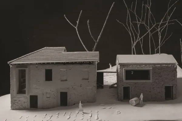 Modell eines architektonischen Projekts mit zwei Gebäuden, umgeben von kahlen Bäumen. Das linke Gebäude hat ein geneigtes Dach, während das rechte ein Flachdach aufweist. Kleine Figuren und Fußspuren sind auf dem Boden sichtbar, was eine menschliche Präsenz andeutet.