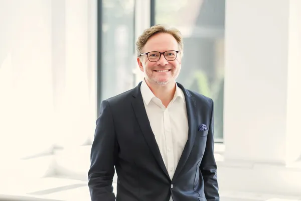 Ein lächelnder Mann in einem Anzug steht vor einem hellen Fenster.