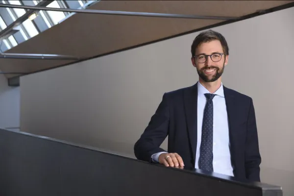 Ein Mann in Anzug und Krawatte steht lächelnd in einem modernen Gebäude mit großen Fenstern.