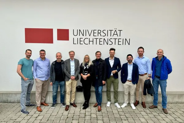 Gruppe von zehn Personen steht lächelnd vor einer Wand mit dem Logo der Universität Liechtenstein. Sie tragen unterschiedliche Freizeit- und Businesskleidung.