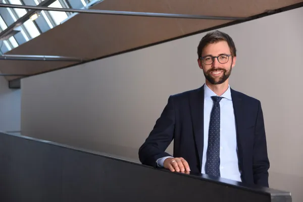 Ein Mann in Anzug und Krawatte steht lächelnd in einem modernen Gebäude mit schrägen Decken und großen Fenstern.