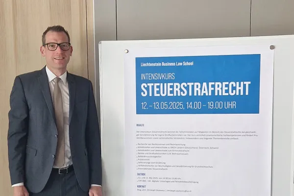 Ein Mann in Anzug steht neben einem Plakat, das den Intensivkurs „Steuerstrafrecht“ der Universität Liechtenstein ankündigt. Das Plakat enthält Informationen zu Datum, Uhrzeit und Inhalten des Kurses.