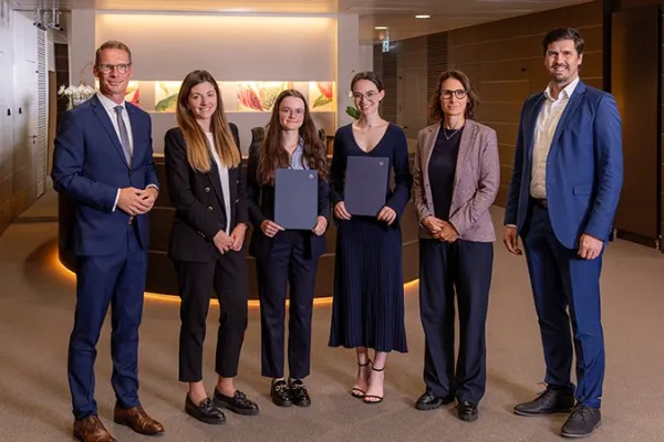Eine Gruppe von sechs Personen steht in einem modernen Bürogebäude. Zwei Frauen in der Mitte halten Zertifikate und lächeln. Sie werden von vier weiteren Personen flankiert, die ebenfalls lächeln. Die Szene vermittelt eine feierliche Atmosphäre, möglicherweise im Rahmen einer Stipendienverleihung an der Universität Liechtenstein.