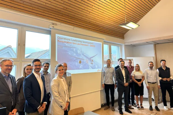 Eine Gruppe von Menschen steht lächelnd in einem Seminarraum vor einer Leinwand, die eine Präsentation zum Operations Leadership Development-Programm der Universität Liechtenstein und SWISS zeigt.