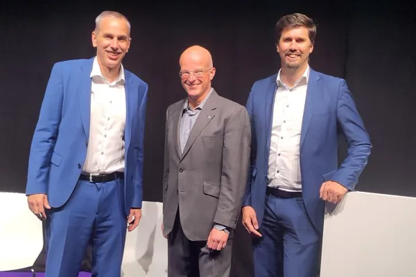 Drei Männer in Anzügen stehen auf einer Bühne mit lila Teppich. Sie lächeln in die Kamera. Im Hintergrund sind schwarze Vorhänge zu sehen.