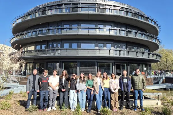 Gruppe von Studierenden und einer Lehrperson vor einem modernen, runden Universitätsgebäude bei sonnigem Wetter.