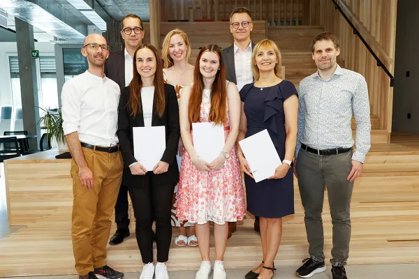 Eine Gruppe von acht Personen steht lächelnd in einem modernen Innenraum. Drei Personen in der vorderen Reihe halten Zertifikate in den Händen. Die Umgebung wirkt hell und einladend mit Holzelementen und Pflanzen.
