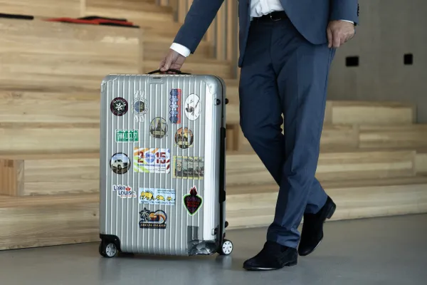 Mann im Anzug zieht silbernen Koffer mit Reiseaufklebern über Campusboden.