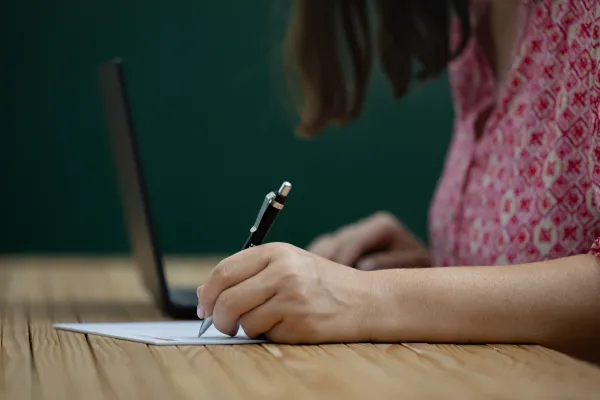 Person schreibt mit Stift auf Papier vor Laptop im Arbeitsraum der Universität Liechtenstein.