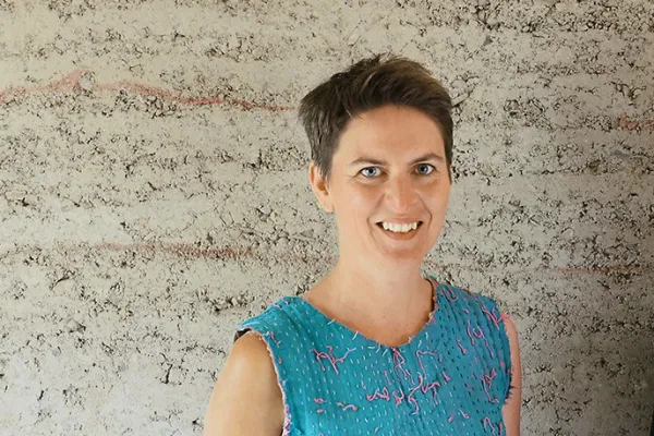Eine lächelnde Person steht vor einer strukturierten Lehmwand. Sie trägt ein blaues Oberteil mit rosa Akzenten.