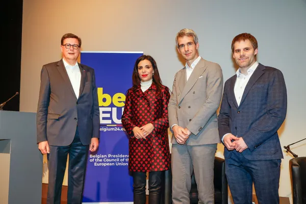 Vier Personen stehen nebeneinander vor einem Banner mit der Aufschrift "Belgian Presidency of the Council of the European Union". Sie tragen formelle Kleidung und blicken in die Kamera.