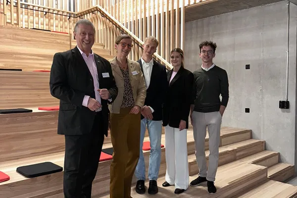 Fünf Personen stehen lächelnd in einer modernen Aula mit Holzstufen und vertikalen Holzlamellen im Hintergrund. Sie tragen formelle Kleidung und posieren für ein Gruppenfoto.