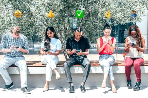 Fünf Personen sitzen auf einer Bank im Freien und schauen auf ihre Smartphones. Über ihren Köpfen sind verschiedene Emojis zu sehen, die Emotionen und Gesten darstellen. Im Hintergrund sind Bäume und ein Gebäude sichtbar.