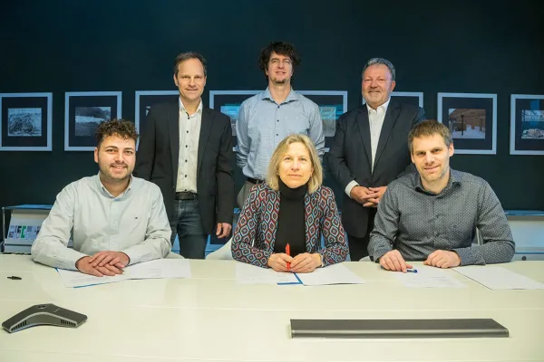 Sechs Personen bei der Unterzeichnung einer Vereinbarung an einem Tisch. Drei Personen sitzen vorne mit Dokumenten, während drei weitere im Hintergrund stehen. Die Szene findet in einem Büro mit Bildern an der Wand statt.