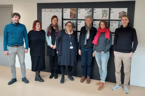 Gruppe von sieben Personen steht vor einer Wand mit dem Titel "PROBONO" und mehreren Plakaten. Sie lächeln in die Kamera und repräsentieren das Service Learning Netzwerk an der Universität Liechtenstein.