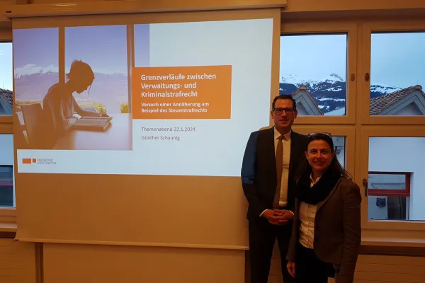Ein Mann und eine Frau stehen vor einer Präsentationsleinwand mit dem Titel "Grenzverläufe zwischen Verwaltungs- und Kriminalstrafrecht" im Seminarraum der Universität Liechtenstein. Im Hintergrund sind schneebedeckte Berge durch die Fenster sichtbar.