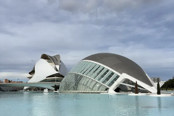Moderne, futuristische Gebäude mit geschwungenen Glas- und Betonformen an einem großen Wasserbecken unter bewölktem Himmel.