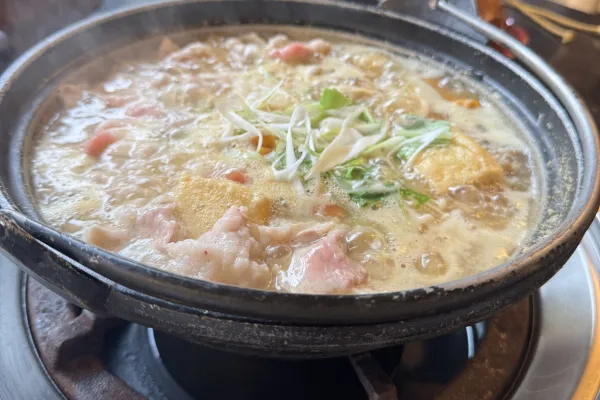 Dampfender Hot Pot mit Fleisch, Tofu und Gemüse in heller Brühe auf einem Tisch-Gasbrenner.